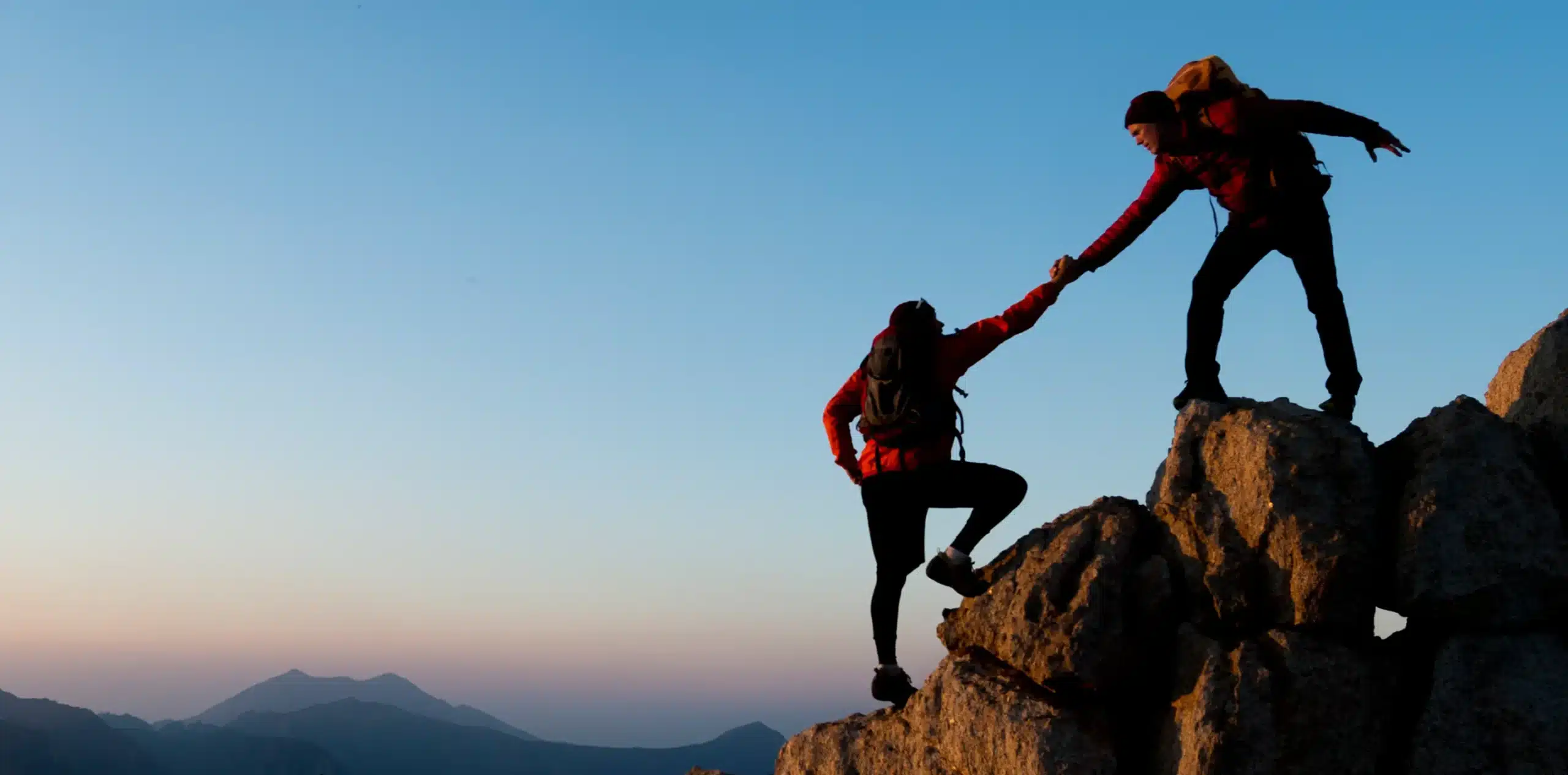 Two hikers silhouetted against a twilight sky as one reaches down to pull another up